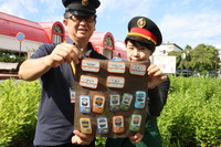 飯田駅を背景に缶バッジをお披露目する尾沢さん（左）ら