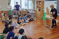 長さの違う3本のスポンジを使った実験装置を、リズムを変えながら揺らすDr.ナダレンジャー（右端）。揺れやすいリズムを受けた建物が大きく揺れる「共振」をこども記者たちに説明しました＝８月２日、諏訪市